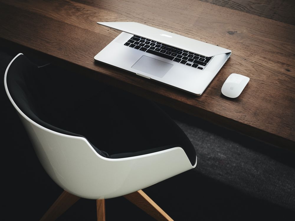 An open laptop and mouse on a dark timber desk beside a pale designer chair.