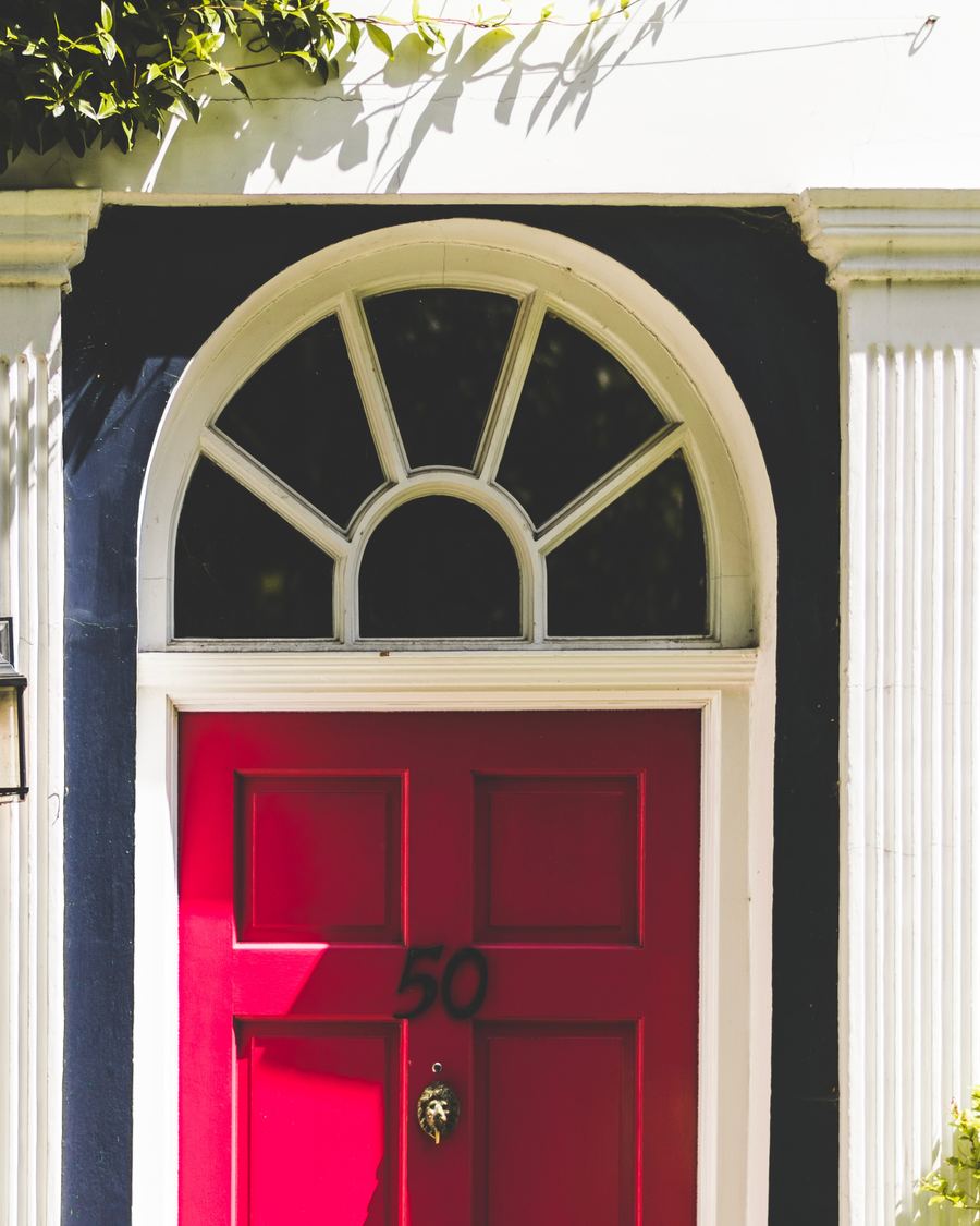 The entrance to a Prime Central London townhouse: a panelled front door beneath a fanlight, framed by fluted white pilasters, with a lit carriage lantern and jasmine growing over the doorway.