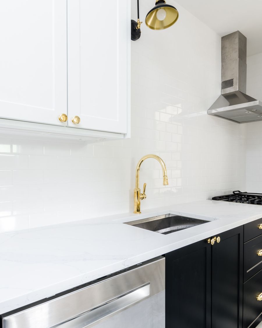 A close view of a kitchen worktop with a brass tap, white metro tiling, a brass wall light and dark cabinetry.