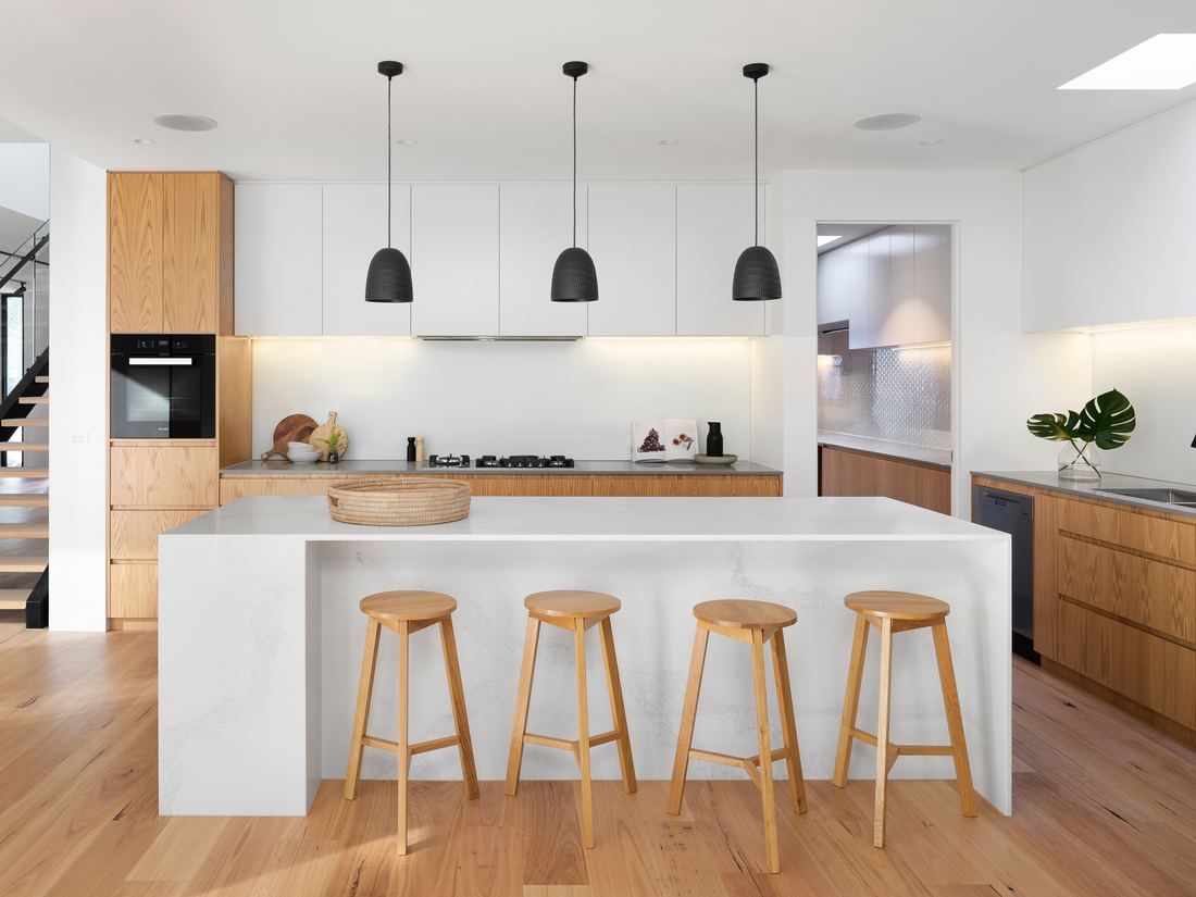 A bright open-plan kitchen with a pale stone island, oak cabinetry, wooden stools and pendant lighting.