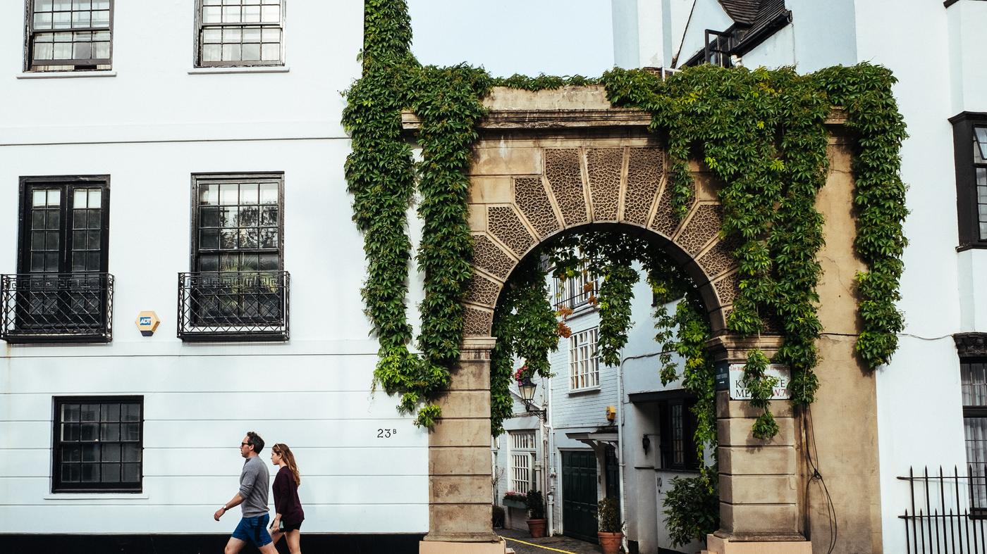 An ivy-covered stone archway leading into a Kensington mews, framed by white stucco houses with black iron balconies.