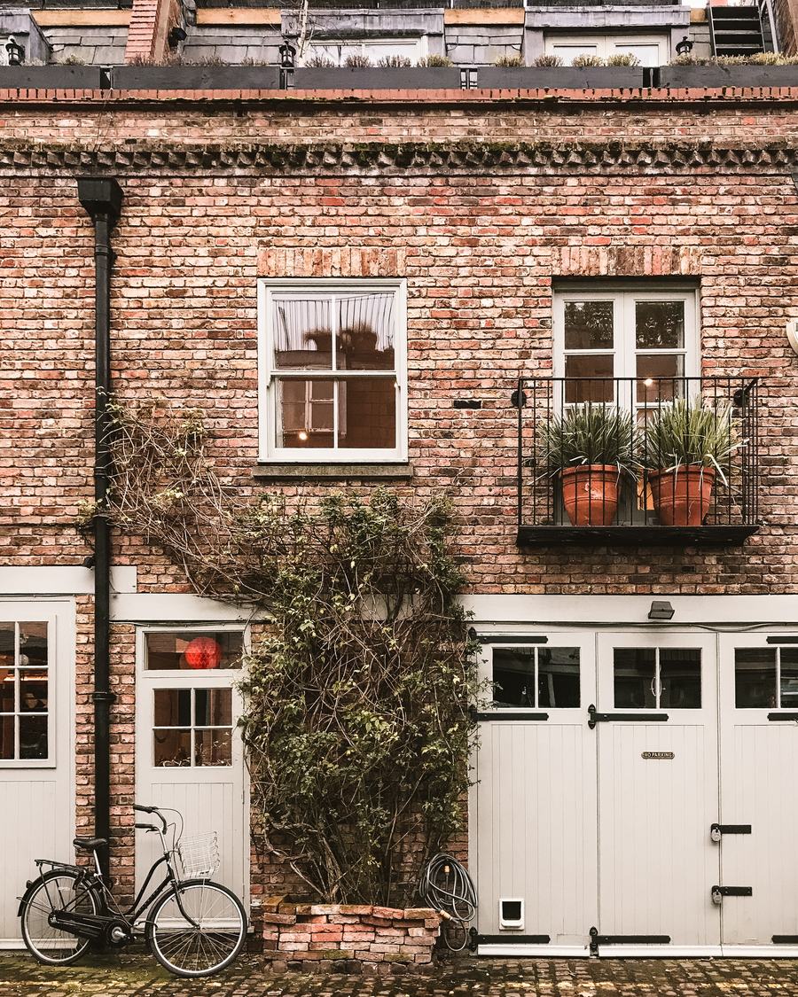 A London brick mews house with white garage doors, a climbing plant and a bicycle resting against the wall.