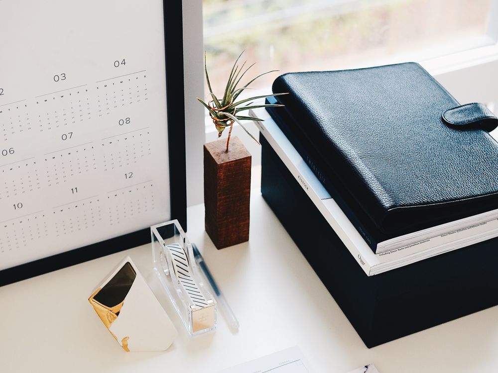 A desk with a monthly planner, notebooks, a leather folder and a framed year calendar beside a window.