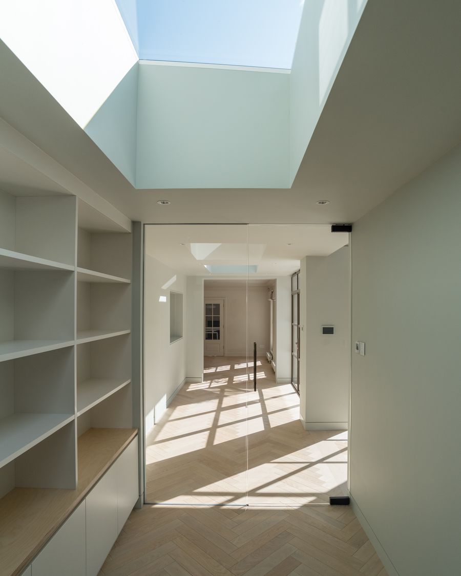 A renovated London hallway with a long roof light, herringbone timber floor, built-in white joinery and a glass door.